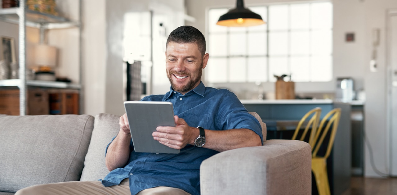 Smiling man relaxing on a couch using a tablet in a bright modern living room
